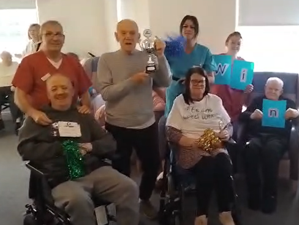 A group of people are sat and stood celebrating a win. The man in the middle is holding up a trophy.